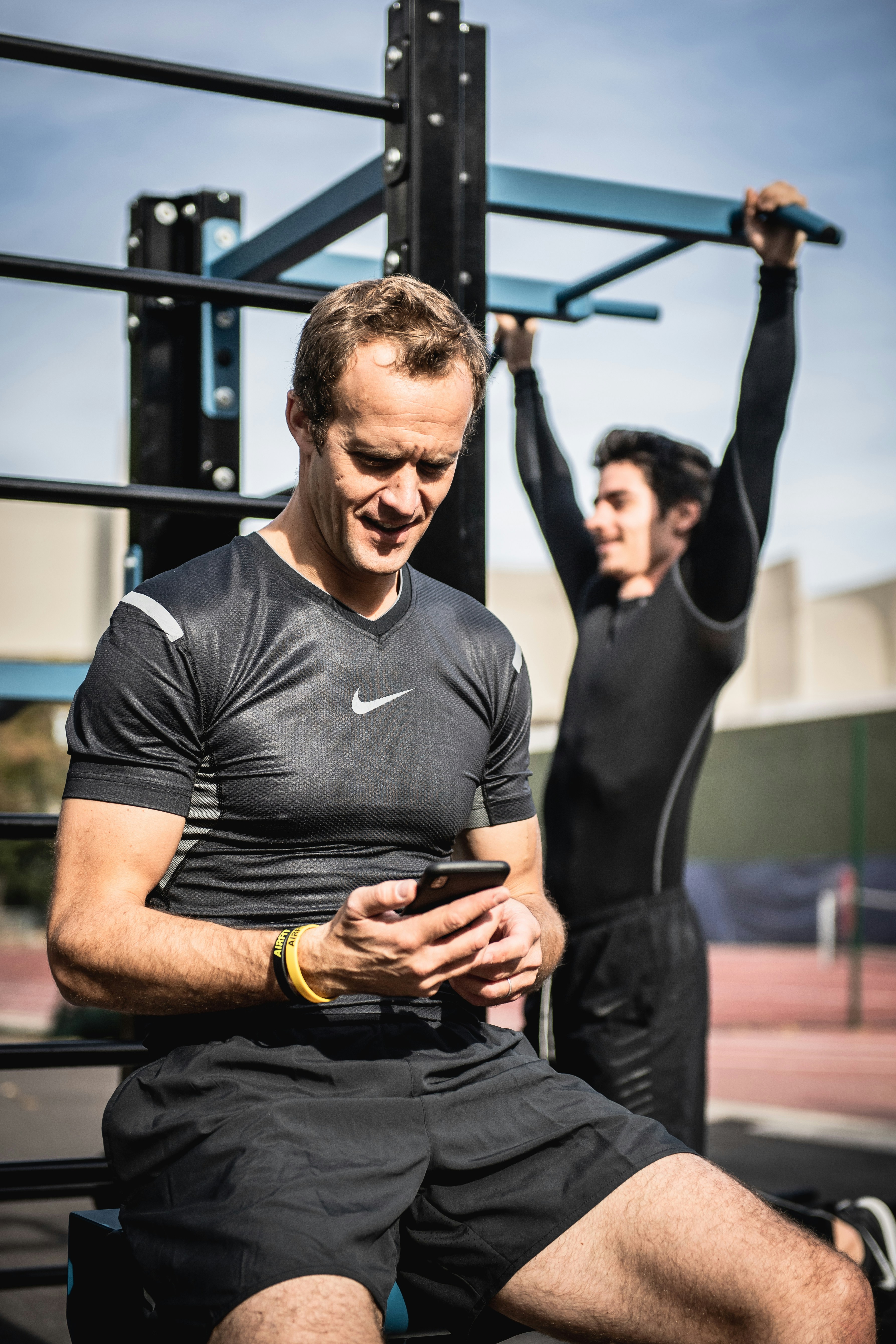 Personal Trainer mit Smartphone an einer Outdoor-Fitnessanlage, im Hintergrund ein Sportler am Klimmzug